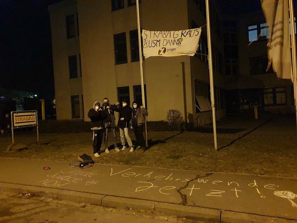 Das Transparent mit dem Slogan "Strabag raus aus'm Danni" hängt zwischen zwei Fahnenstangen, daneben eine Gruppe von jungen Attacis, die schwarz gekleidet sind und kämpferisch aussehen.