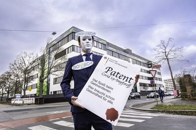 Ein Mensch im Anzug und Biontech-Logo am Revers hält eine übergroße Patenturkunde auf Corona-Impfungen. Im Hintergrund steht die Biontech-Zentrale