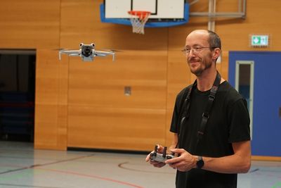 In der Turnhalle steht ein Drohnenfotograf, neben ihm schwebt seine Drohne