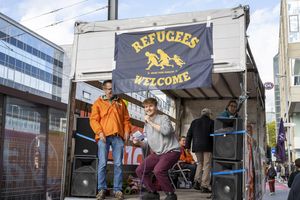 Ein junger Mann steht auf dem Lautsprecherwagen und spricht ins Mikrophon. Neben ihm steht ein älterer Mann in oranger Jacke und hinter den beiden ist eine Refugees-Welcome-Flagge zu sehen
