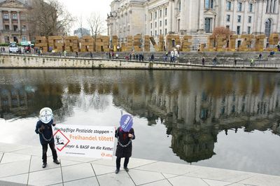 Zwei Attac-Aktivist*innen die eine Maske mit den Logos von Bayer und BASF tragen halten ein Transparent mit der Aufschrift "Hochgiftige Pestizide stoppen – Freihandel tötet – Bayer und BASF an die Kette!" im Hintergrund, hinter der Spree stehen vor dem Reichstag aus Strohballen geformt die Buchstaben "AGRARWENDE JETZT"
