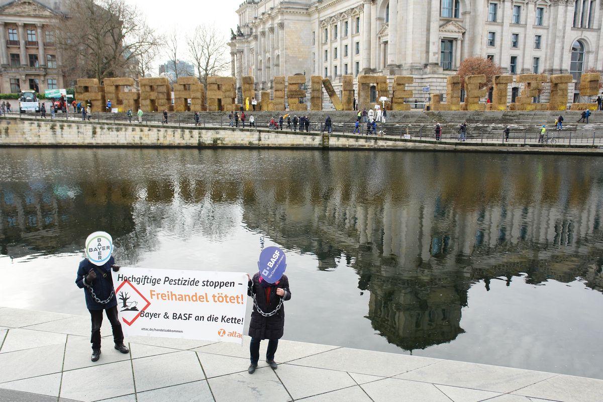 Zwei Attac-Aktivist*innen die eine Maske mit den Logos von Bayer und BASF tragen halten ein Transparent mit der Aufschrift "Hochgiftige Pestizide stoppen – Freihandel tötet – Bayer und BASF an die Kette!" im Hintergrund, hinter der Spree stehen vor dem Reichstag aus Strohballen geformt die Buchstaben "AGRARWENDE JETZT"