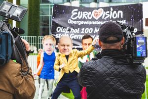 Im Vordergrund stehen zwei Kameramänner, die im Hintergrund schrill-bunt verkleidete Menschen mit Masken von Boris Johnson und Olaf Scholz aufnehmen, ganz hinten ist unscharf ein Transparent zu erkennen.
