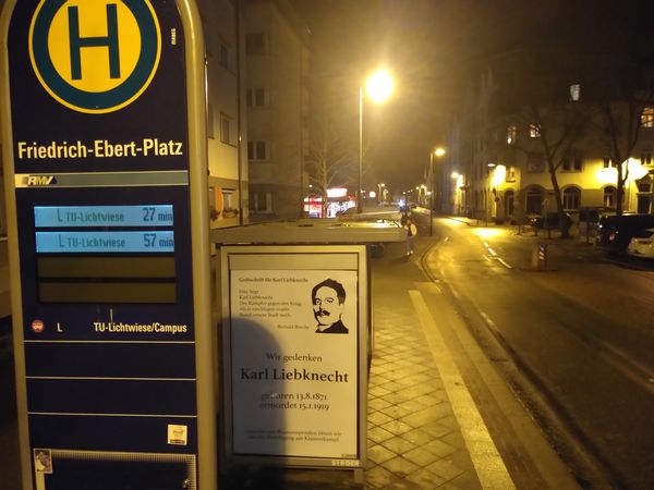 In der Bushaltestelle Friedrich-Ebert-Platz hängt ein Geadbustetes Plakat im Stil einer Traueranzeige mit dem Text "Grabinschrift für Karl Liebknecht. Hier liegt/Karl Liebknecht/Der Kämpfer gegen den Krieg/Als er erschlagen wurde/Stand unsere Stadt noch. Bertold Brecht. Wir Gedenken Karl Liebknecht geboren 13.8.1871 ermordet 15.1.1919. Anstelle von Blumenspenden bitten wir um die Beteiligung am Klassenkampf"