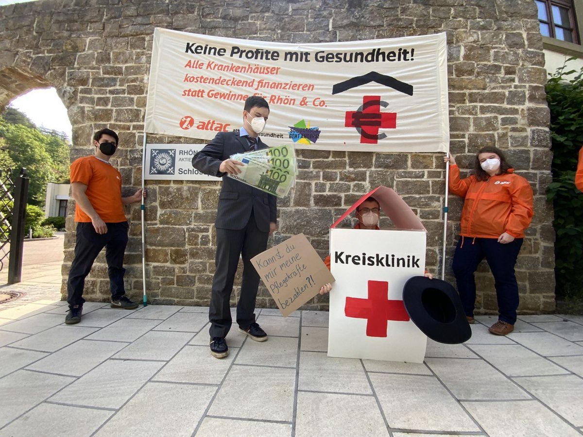 Ein Man im Anzug und Klinikkonzernlogos steht neben einem bettelnden Menschen in einem Krankenhauskostüm. Dahinter ein Transparent "Keine Profite Mit Gesundheit"