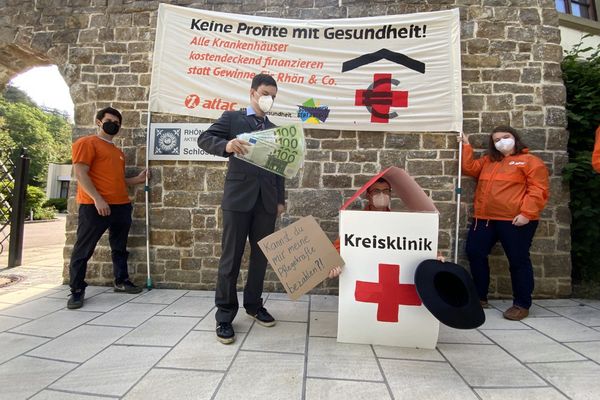 Bild: Stephan Kettner Ein Man im Anzug mit Logos privater Klinikkonzerne und Geldscheinen steht neben einem Menschen im Kostüm eine Kreisklinik, die Bettelt, im Hintergrund das Aktionstransparent.