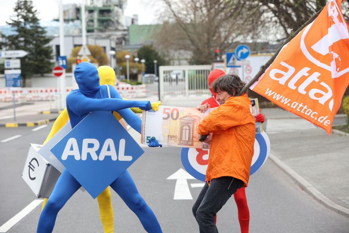 Menschen in farbigen Ganzkörperanzügen mit den Logos von Aral, Shell und Esso ziehen an einem Geldschein auf der anderen Seite ziehen Attacies.
