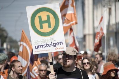 Ein Aktivist hält ein Schild mit einem Haltestellenzeichen und dem Logo der Attac-Kampagne "einfach.umsteigen", hinter ihm läuft der Attac-Block