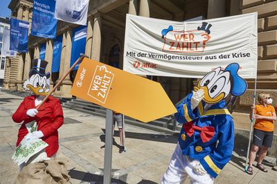 Dagobert zeigt mit einem Pfeil auf dem "Wer zahlt?" steht auf den erschrocken schauenden Donald, im Hintergrund das Transparent mit der Aufschrift "Wer Zahlt? Mit der Vermögenssteuer aus der Krise"