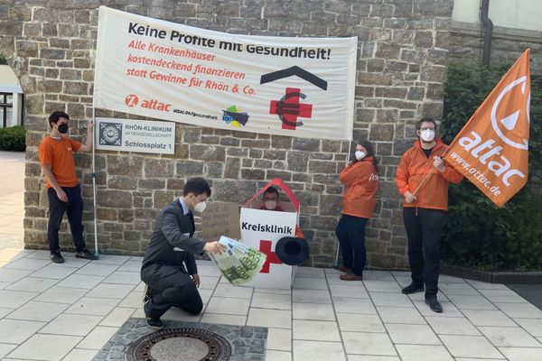 Bild: Stephan Kettner Ein Man im Anzug mit Logos privater Klinikkonzerne und Geldscheinen steht neben einem Menschen im Kostüm eine Kreisklinik, die Bettelt, im Hintergrund das Aktionstransparent und ein Mensch mit Attac-Fahne