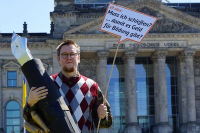 Ein Aktivist hat eine Camouflage-Uniform an und trägt darüber einen Pullunder sowie eine Fliege, in seiner Hand hält er einen überdimensionierten Füller mit einem ebenfalls überdimensionierten Patronengurt. In der anderen Hand hält er ein Schild mit der Aufschrift "Muss ich schießen, damit es Geld für Bildung gibt?"