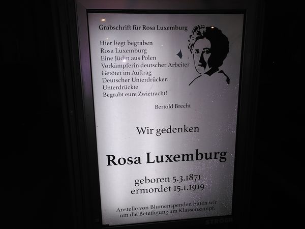In einer Bushaltestelle hängt ein Geadbustetes Plakat im Stil einer Traueranzeige mit dem Text "Grabinschrift für Rosa Luxemburg. Hier liegt begraben/Rosa Luxemburg/Eine Jüdin aus Polen/Vorkämpferin deutscher Arbeiter/Getötet im Auftrag/Deutscher Unterdrücker./Unterdrückte/Begrabt eure Zwietracht! Bertold Brecht. Wir gedenken Rosa Luxemburg. geboren 5.3.1871 ermordet 15.1.1919 Anstelle von Blumenspenden bitten wir um die Beteiligung im Klassenkampf.“