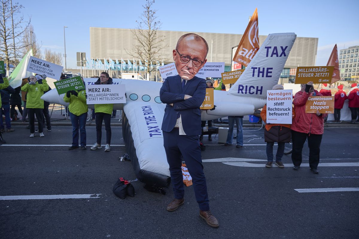 Mensch mit Merz-Maske steht vor einem aufgeblasenen Flugzeug auf dem Tax the Rich steht