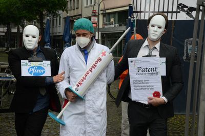 Fotograf + R-Mediabase Ein als Arzt verkleideter Mensch mit einer Spritze, auf der Solidarität steht, wird von zwei Menschen im Anzug, die die Pharmaindustrie symbolisieren abgeführt.