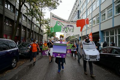 Auf der IAA-Demo läuft ein Mensch in einem Buskostüm und einem grünen Daumen nach oben neben einem Menschen in einem SUV-Kostüm mit Dinomaske mit einem roten Daumen nach unten. Hinter ihnen halten Aktivist*innen ein Transparent mit dem Slogan "Autosaurus ins Museum"