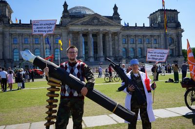 Vor dem Reichstag stehen zwei verkleidete Aktivist*innen, beide haben eine Camouflage-Uniform an. Einer trägt darüber Pullunder und Fliege und hält einen überdimensionierten Füller mit ebenfalls überdimensioniertem Patronengurt, in der anderen Hand hält er eine Sprechblase mit der Aufschrift "Muss ich erst schießen, damit es Geld für Bildung gibt?". Die zweite Aktivistin trägt einen Pflegekittel und OP-Haube über ihrer Uniform und hält ein aufblasbares Maschinengewehr und eine aufblasbare Spritze in der Hand sowie eine Sprechblase mit der Aufschrift "Muss ich schießen, damit es Geld für die Pflege gibt?"