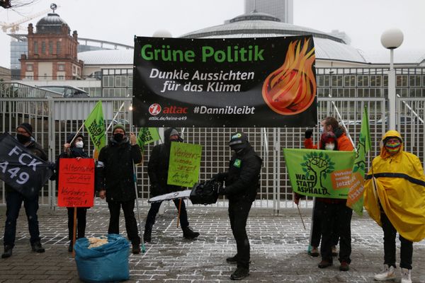 Menschen protestieren vor der Messe, Ein Mensch mit Polizeiuniform auf dem ein Günenlogo ist hält eine Motorsäge in der hand. Im Hintergrund ist ein Banner mit der Aufschrift "Grüne Politik - Dunkle Aussichten für das Klima"