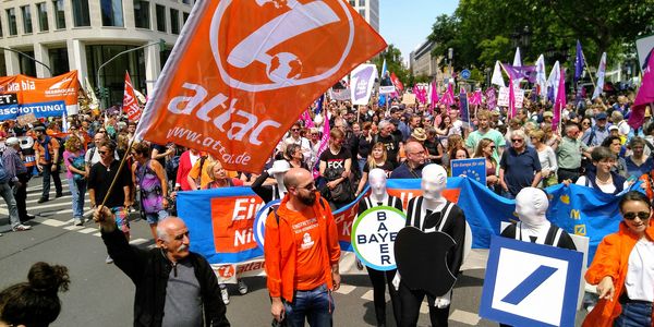 Demobild mit einer großen Attac-Fahne und Menschen die Als Logos verschiedener Großkonzerne verkleidet sind