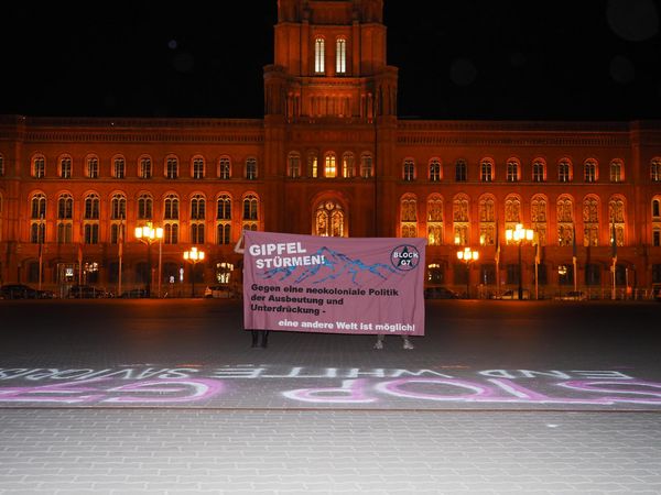 Vor dem nächtlichen Roten Rathaus in Berlin stehen Aktivist*innen die ein Banner zur Mobilisierung gegen den G7-Gipfel in Elmar halten. Vor sich haben sie mit Sprühkreide "Stop G7 – End white Saviorism" geschrieben