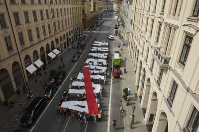 Blick von ca. 20m Höhe auf eine Straße auf dem Menschen mit großen Stoffbuchstaben das Wort "Autobahn" formen, wobei das Auto rot durchgestrichen ist