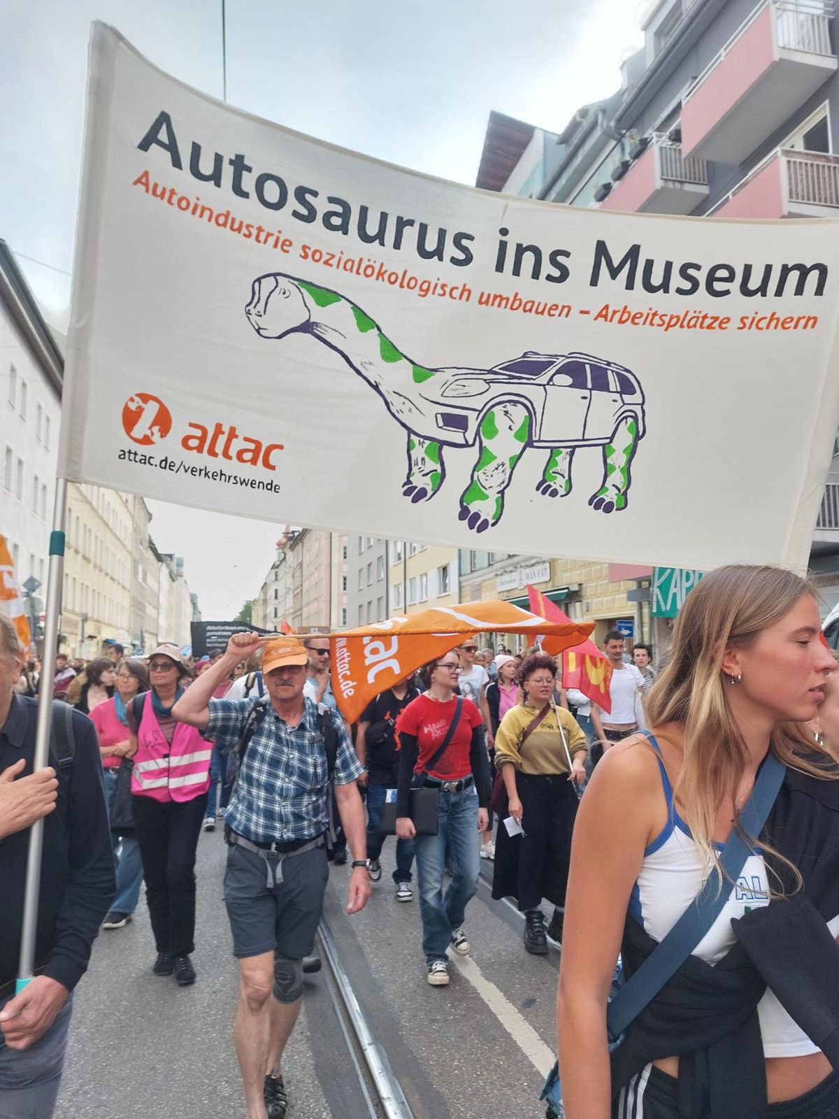Demonstrationszug mit Transparent, darauf ein Dinosaurier mit Autokörper. Auf dem Transparent steht geschrieben "Autosurus ins Museum - Autoindustrie souialökologisch umbauen - Arbeitsplätze sichern" außerdem das attac-logo. Mehrere Menschen tragen eine Attac Fahne.
