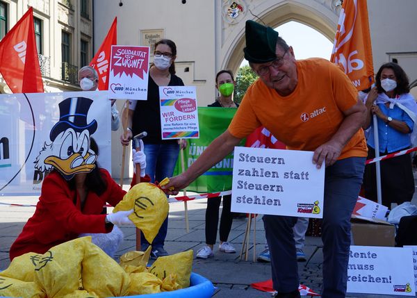Ein Attacie mit Robin Hood-Hut entreißt einer als Dagobert Duck verkleideten Person einen Geldsack. Der Attaci hält ein Schild mit dem Slogan "Steuern zahlen statt Steuern stehlen"