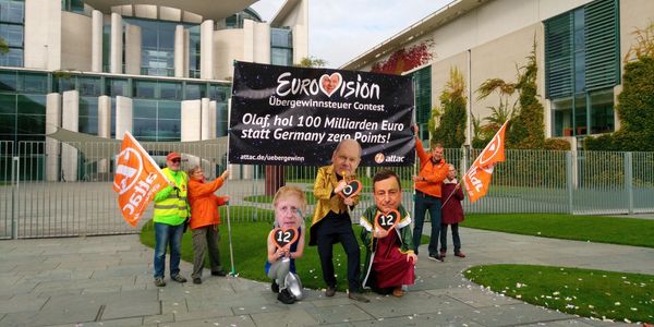 Vor dem Kanzleramt steht eine Gruppe Demonstrant*innen. Sie halten ein Banner mit dem Slogan "Eurovision Übergewinnsteuer Contest. Olaf, hol 100 Milliarden Euro statt Germany Zero Points!" Davor stehen drei schrill und glitzernd verkleidete Aktivist*innen, die Teilnehmer am Contest darstellen mit den Masken von Olaf Scholz, Boris Johnson und Mario Draghi, Olaf Scholz hält 0 Punkte in der Hand, die anderen beiden 12 Punkte