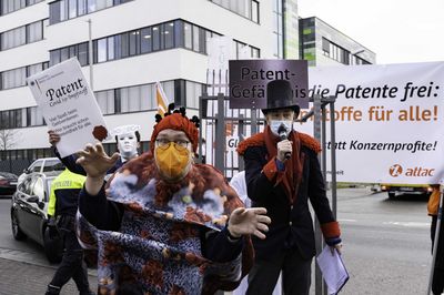 Ein Mensch im Viruskostüm hüpft in erschreckender Mine nach vorne. Im Hintergrund sieht man einen karnevalesk verkleideten Menschen der singt.