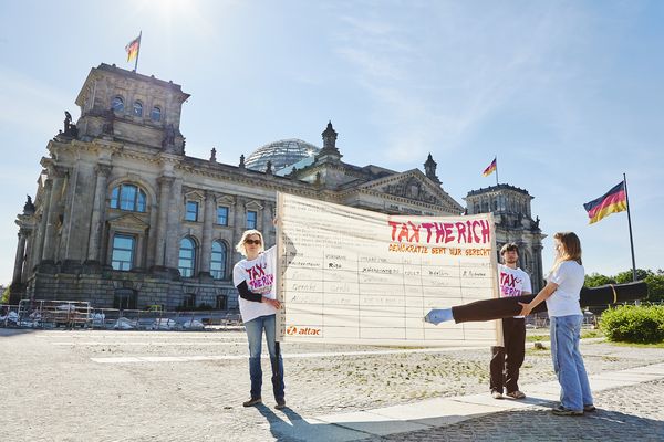Zwei Aktivist*innen halten ein großes Banner auf der eine überdimensionierte Unterschriftenliste für Tax the Rich aufgemalt ist, eine Person unterschreibt mit einem überdimensionierten Füller. Im Hintergrund ist das Reichstagsgebäude zu sehen.
