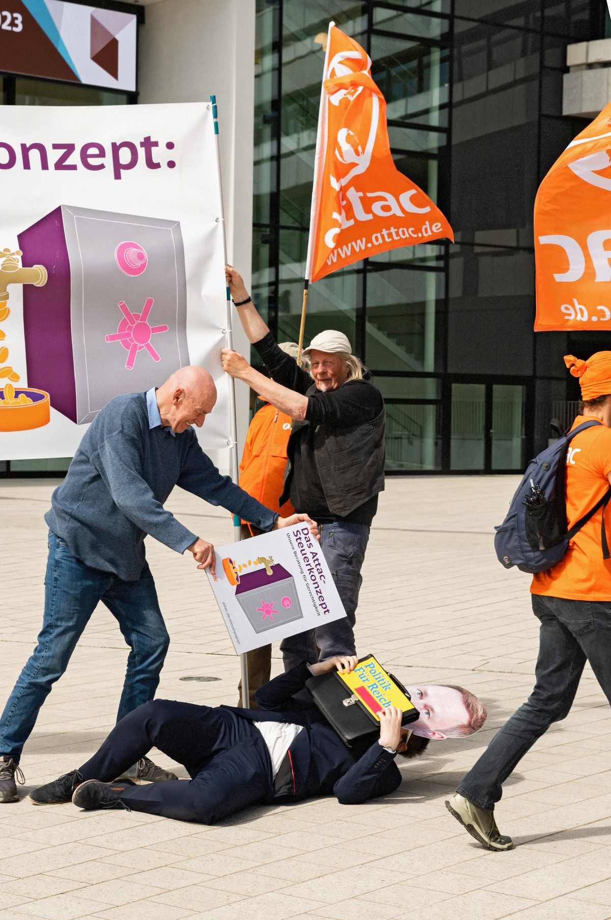 Der Mensch im Christian-Lindner-Kostüm liegt am Boden, darüber hält ein Attac-Aktivist das Attac-Steuerkonzept.